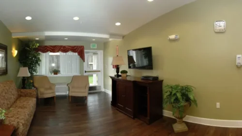 A panoramic shot of a communal seating area with a patterned couch, two armchairs, a television, and a hallway visible on the right
