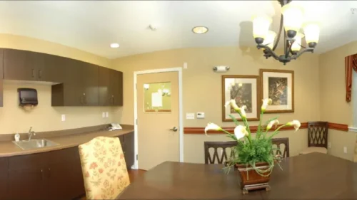 A panoramic shot of a kitchenette with dark wood cabinets, a sink, a dining table with chairs, and a window with a valance