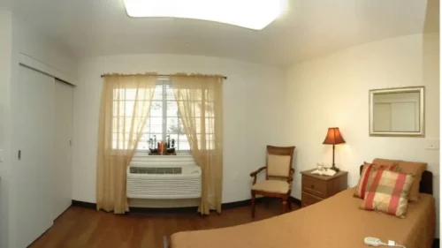 A panoramic view of a bedroom featuring a bed, a dresser with a lamp, and a bathroom vanity with a mirror and sink